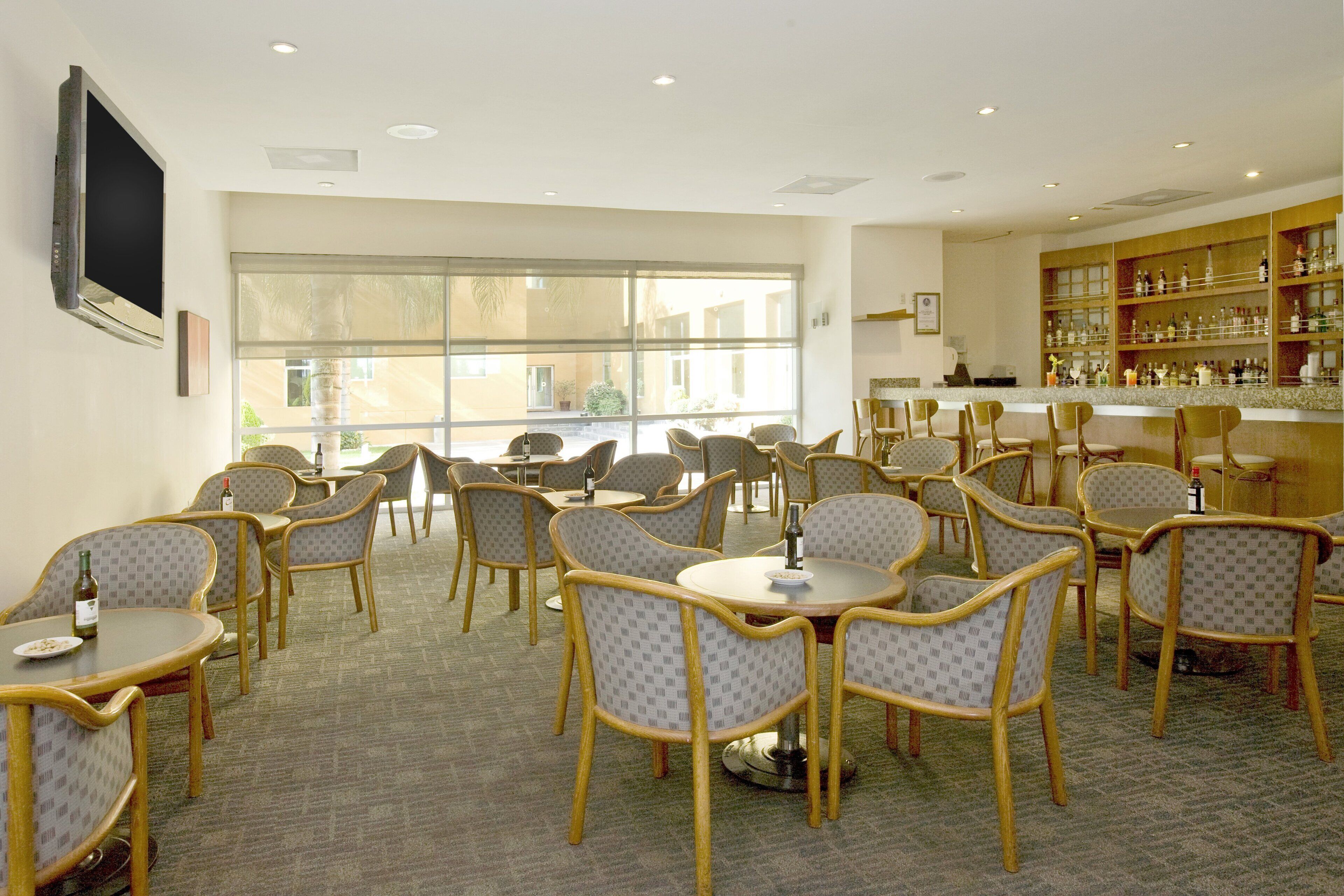A hotel lobby with round tables and chairs, a bar area with shelves of bottles, and large windows providing natural light.