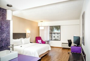 Habitación de hotel con cama, escritorio con computadora, silla, ventana y baño con lavamanos.