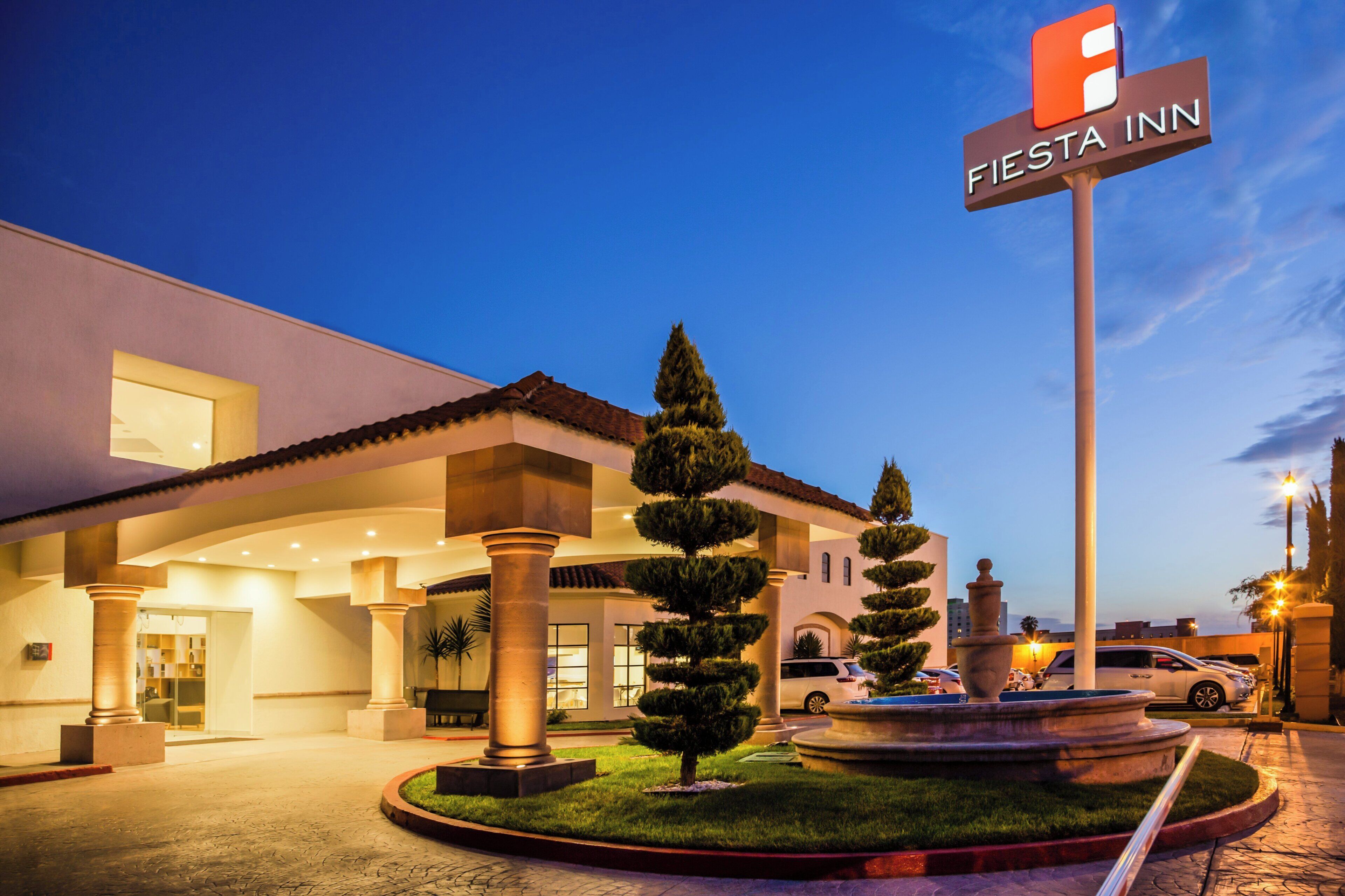 A hotel entrance with a sign reading 'Fiesta Inn,' a well-lit facade, and a landscaped area with a fountain.