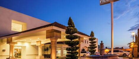 A hotel entrance with a sign reading 'Fiesta Inn,' a well-lit facade, and a landscaped area with a fountain.