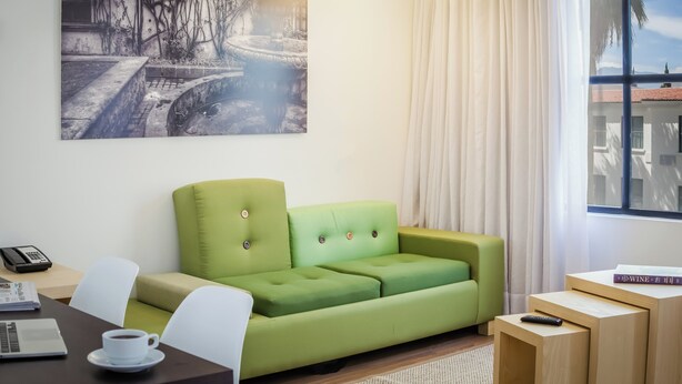 A modern room with a green sofa, a desk with a laptop, a phone, and a remote control. - Fiesta Inn Saltillo (Saltillo)