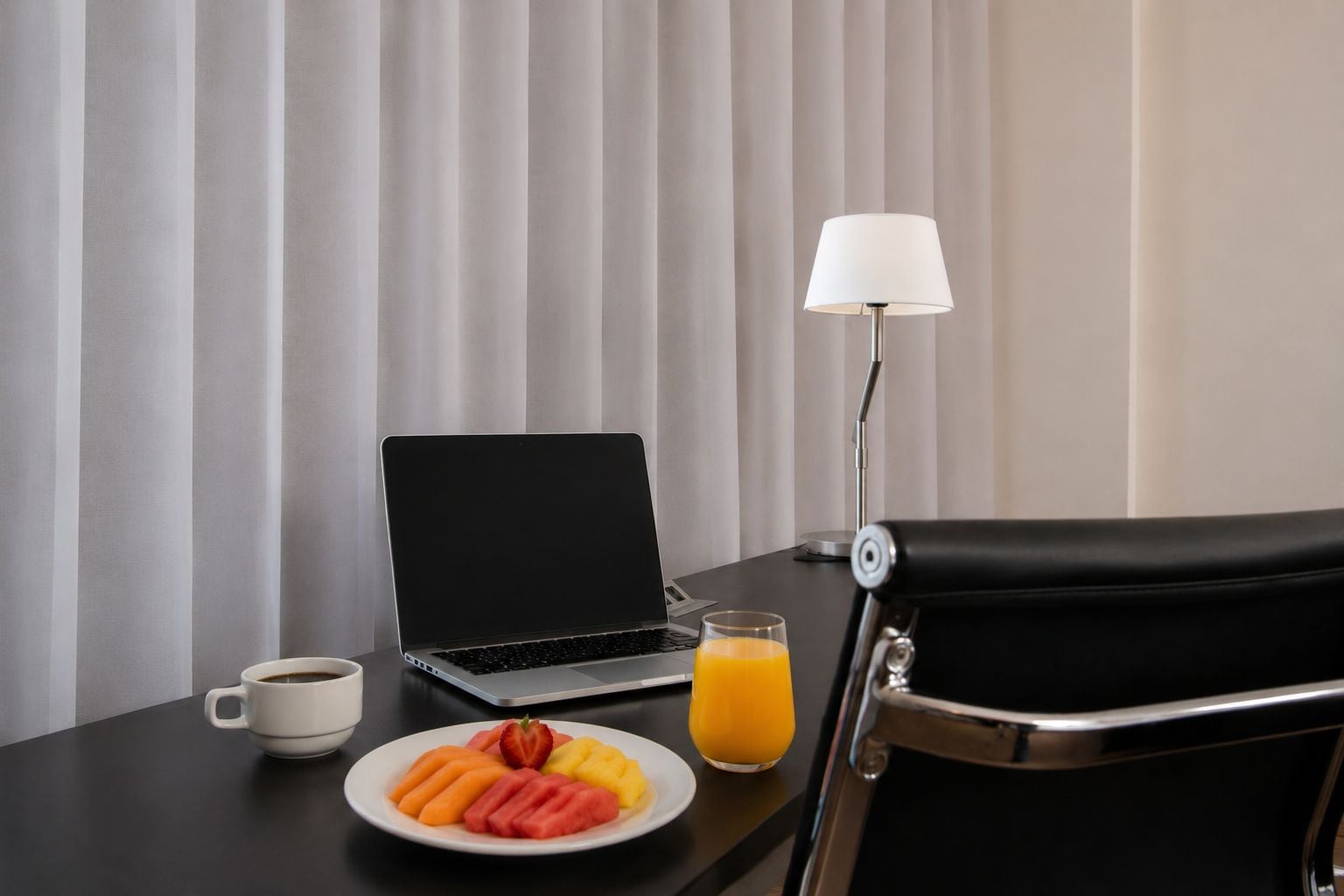 A hotel room with a bed, a desk with a laptop, a coffee cup, a plate of fruit, and a glass of orange juice.