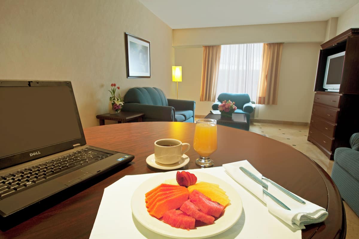 Quarto de hotel com um laptop, uma tábua de frutas, uma xícara de café e um copo de suco de laranja sobre uma mesa.