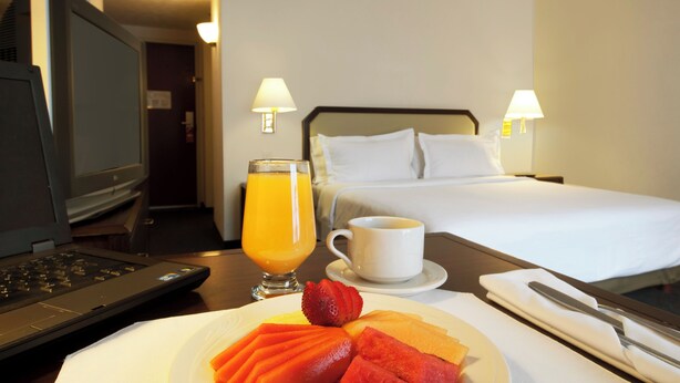 A hotel room with a bed, a desk with a laptop, a plate of fruit, a glass of orange juice, and a cup of coffee. - Fiesta Inn Toluca Tollocan (Toluca)
