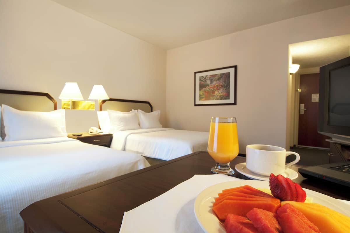 Quarto de hotel com duas camas, uma mesa de jantar com frutas e suco, e uma televisão.