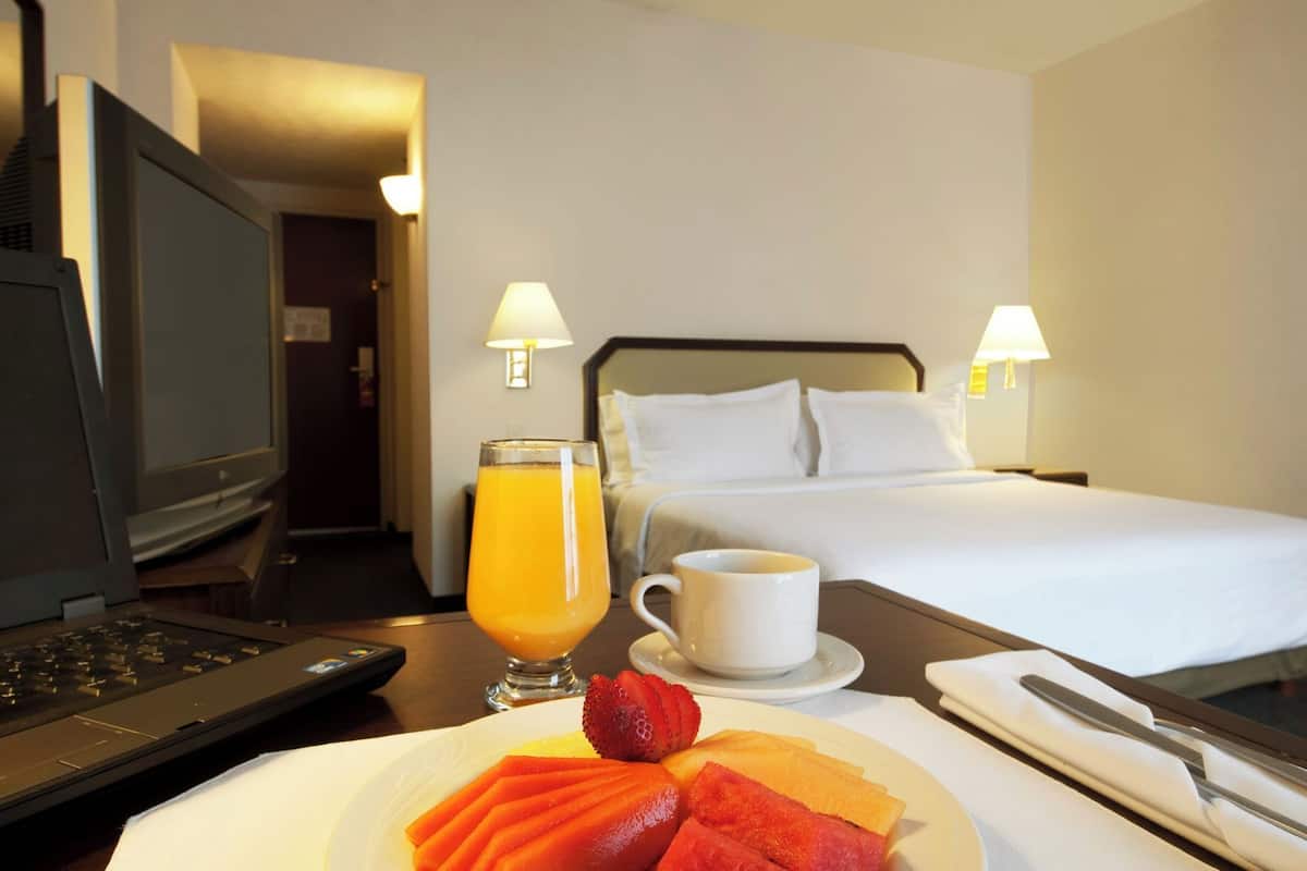 Quarto de hotel com cama, abajures, um laptop, uma tábua de frutas, um copo de suco de laranja e uma xícara de café.
