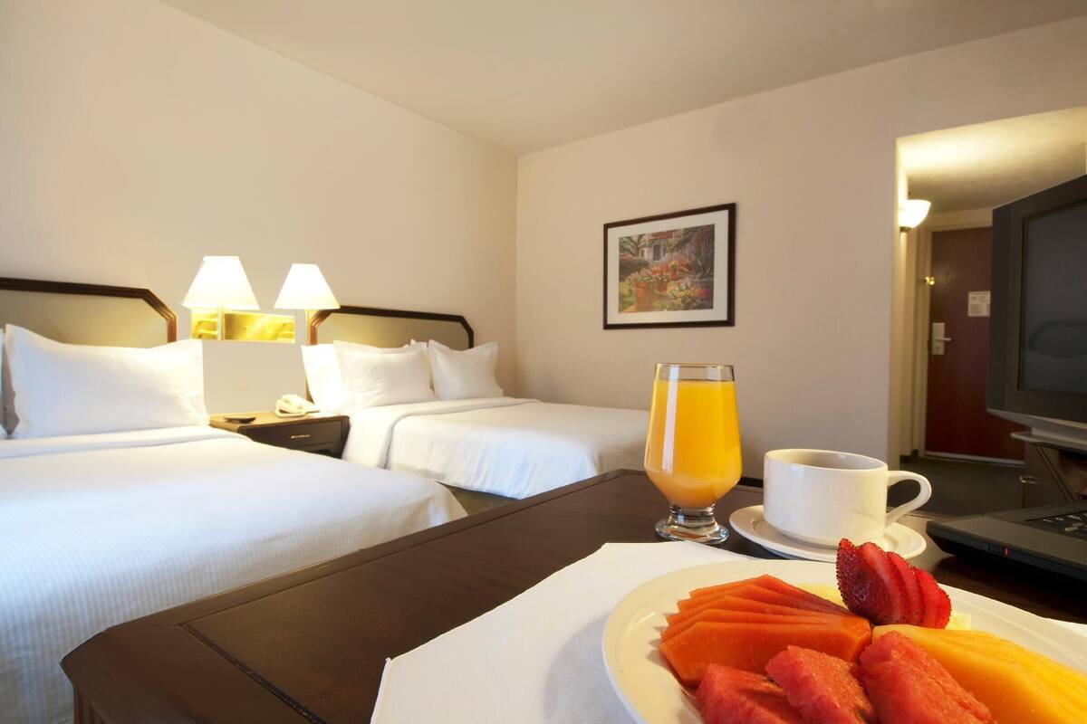 Quarto de hotel com duas camas, uma escrivaninha com computador, uma travessa de fruta, um copo de sumo de laranja e uma chávena de café.