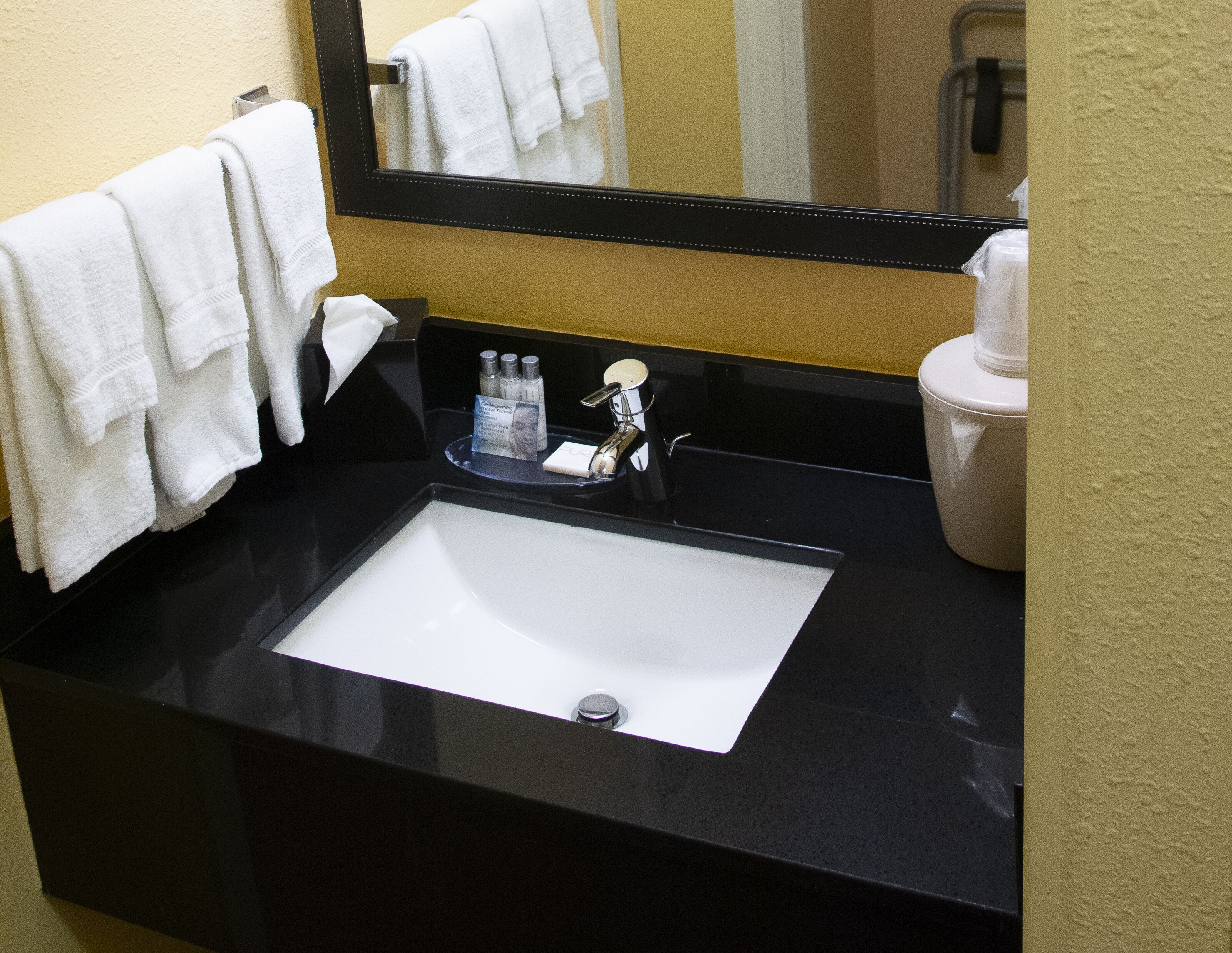 Un baño con encimera negra, un lavabo blanco y un espejo encima. Hay toallas colgadas en la pared y artículos de tocador en la encimera.