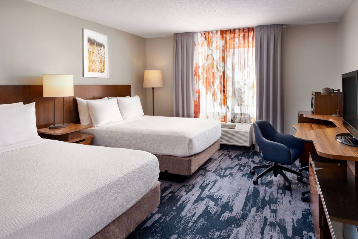 A hotel room with two beds, a desk, a chair, a TV, and a window with curtains.