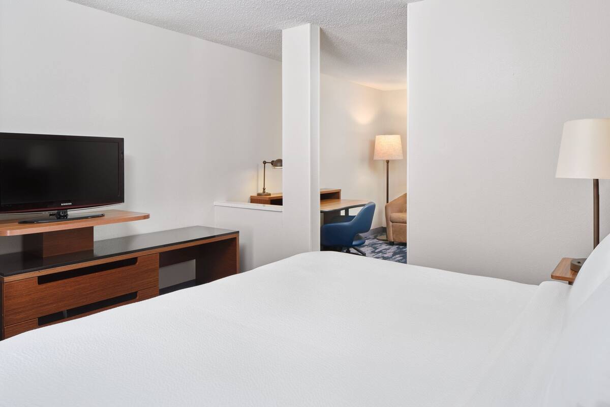A hotel room with a bed, a TV on a wooden stand, a desk with a lamp, and a chair.