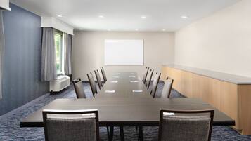 Un salón de conferencias con una mesa larga, sillas, una pantalla de proyector y una pequeña zona de kitchenette.