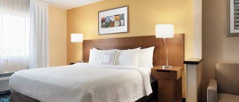 A hotel room with a large bed, two bedside lamps, a headboard, a framed picture on the wall, and a chair.
