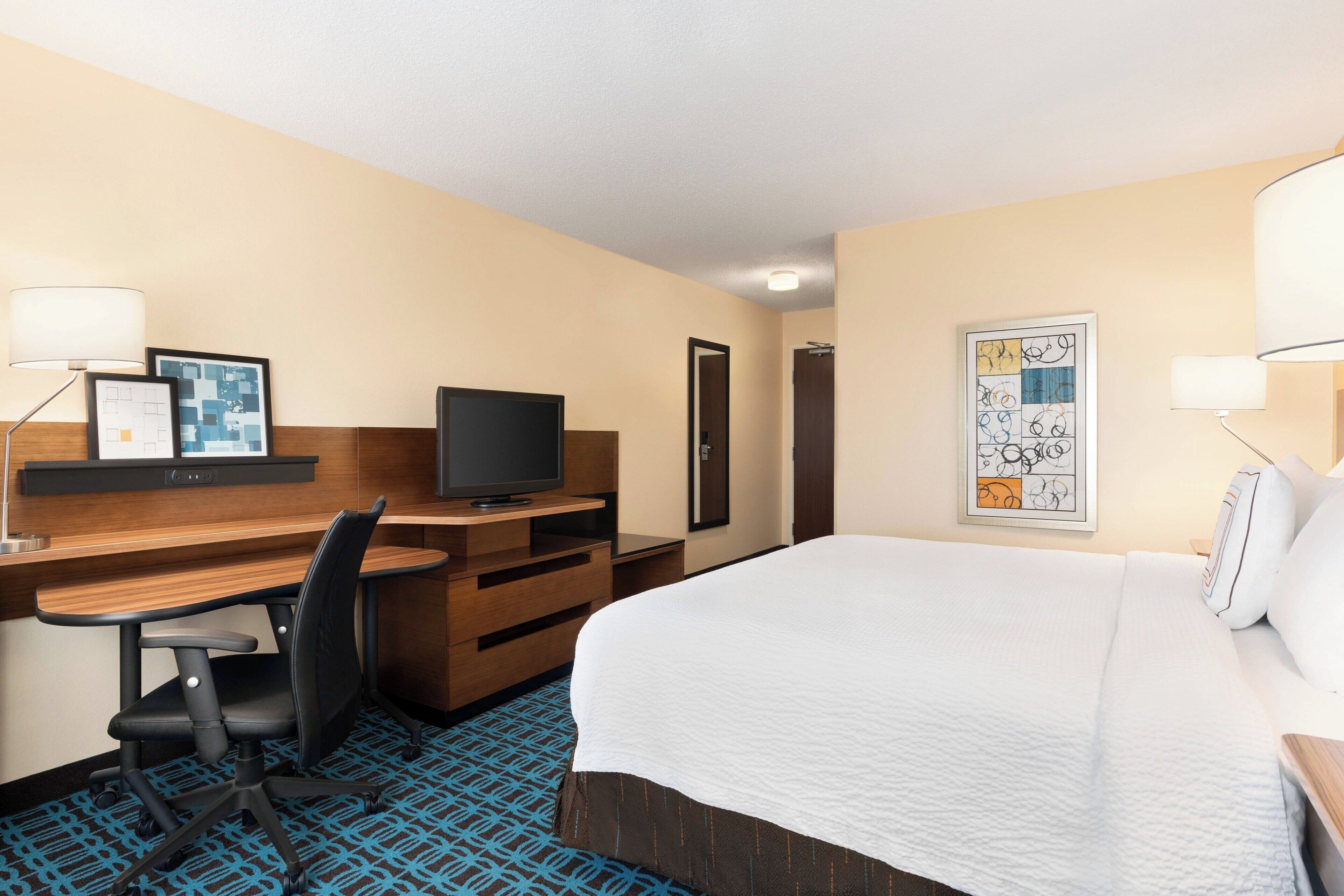 A hotel room with a bed, desk, chair, television, and artwork on the walls.