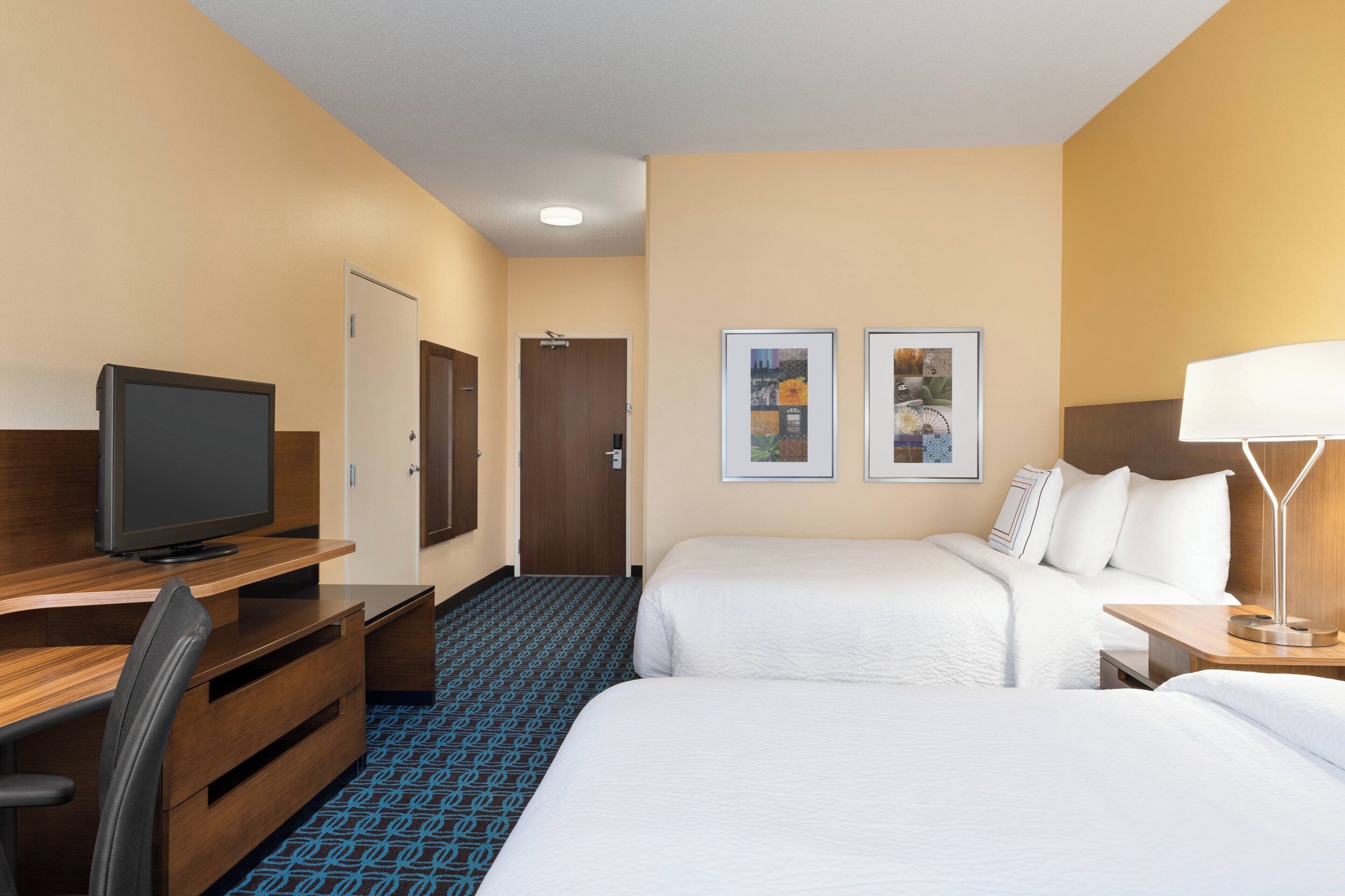A hotel room with two beds, a desk with a TV, a door, and two framed pictures on the wall.