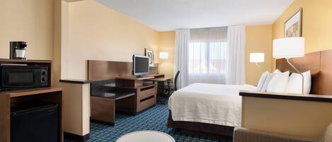 Habitación de hotel con cama, escritorio con computadora, microondas, cafetera, televisión y refrigerador pequeño.