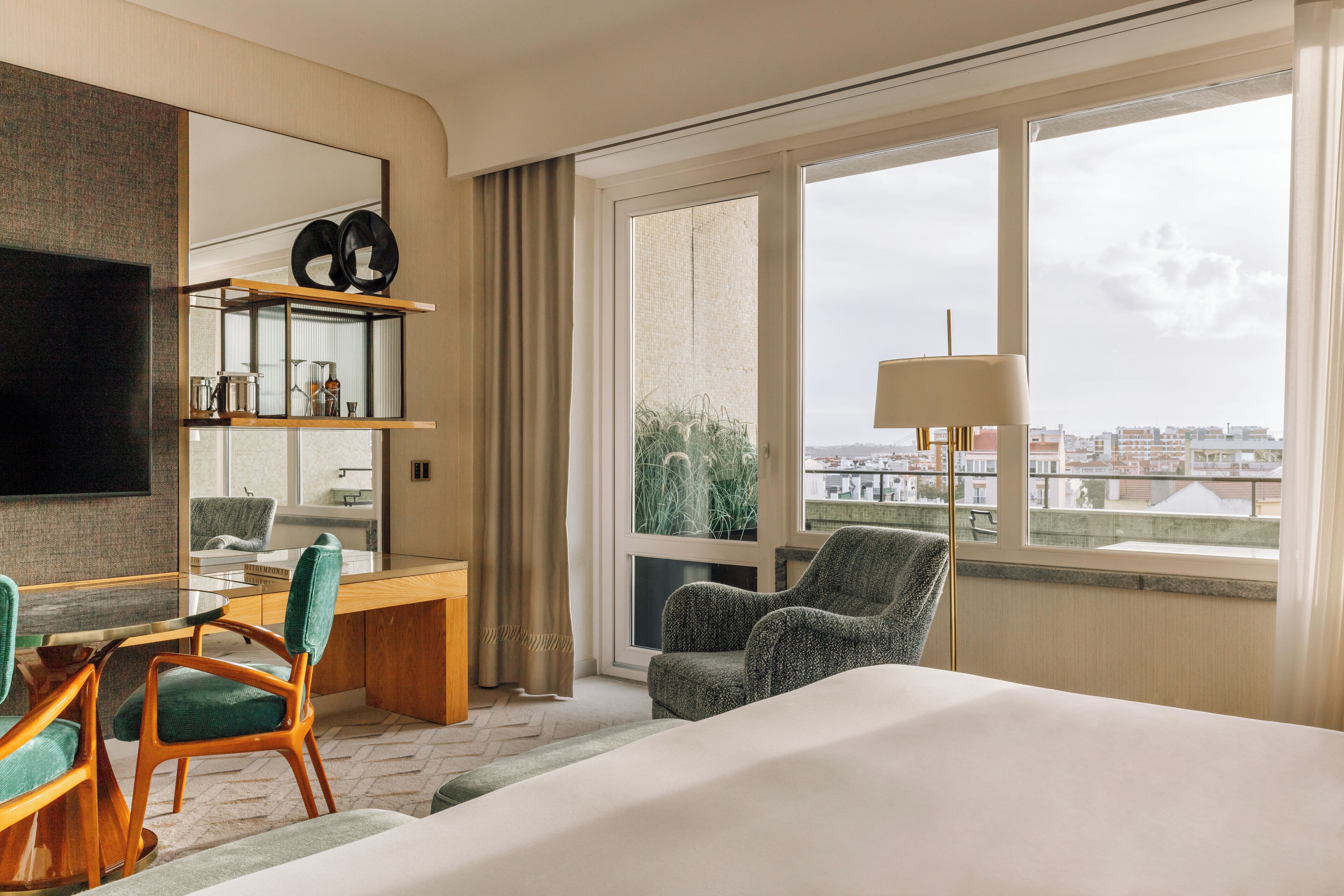 Habitación de hotel con cama, escritorio, dos sillas, televisor, ventana con vista a edificios y lámpara.