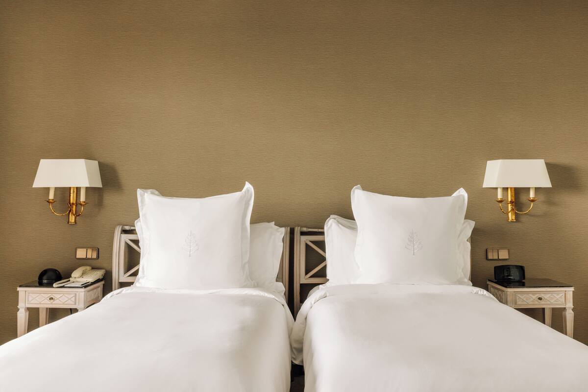 A hotel room with two beds, a nightstand with a phone and lamp, and a wall-mounted lamp.
