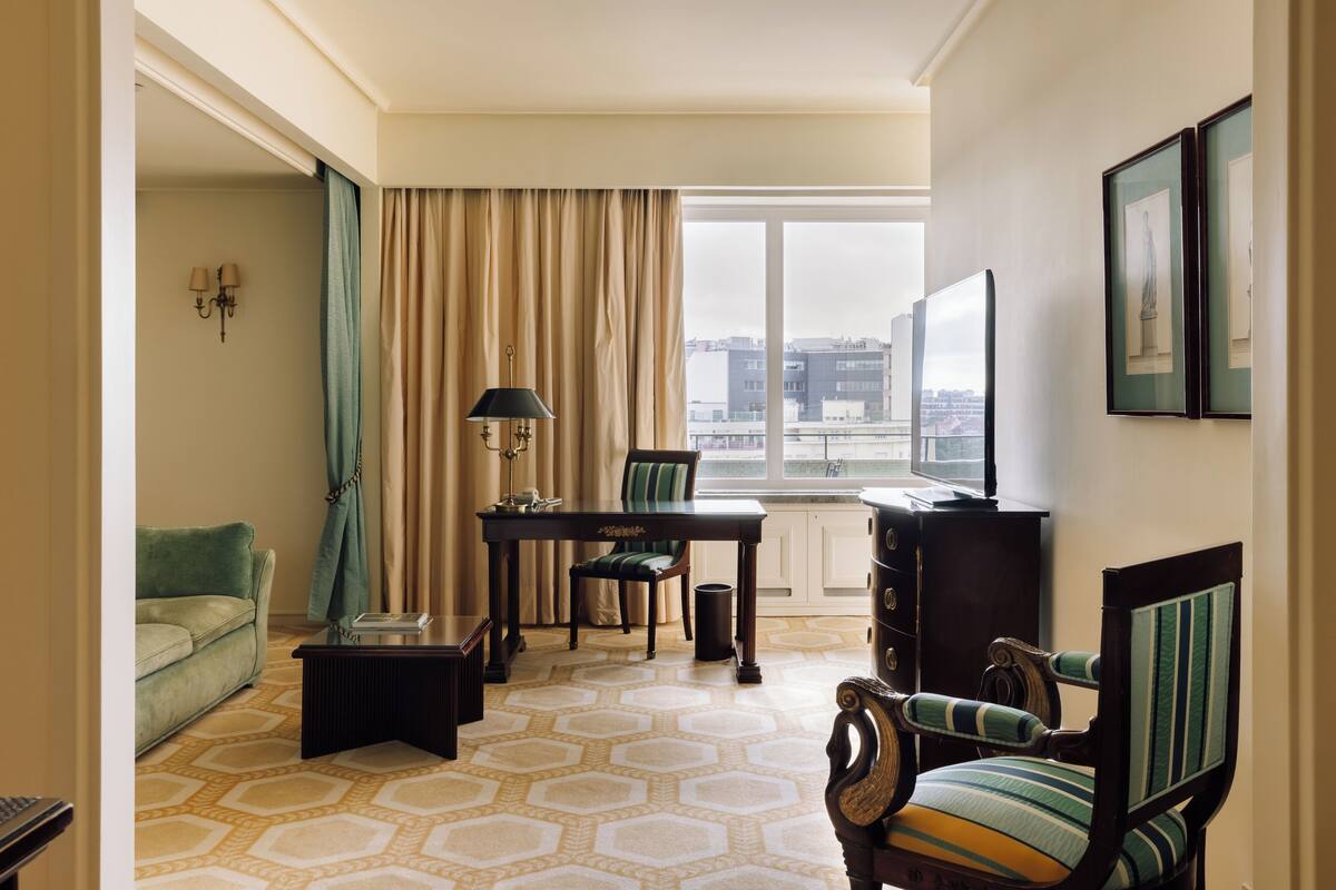 A hotel room with a sofa, a dining table with chairs, a lamp, a TV, and a view of the city through large windows.