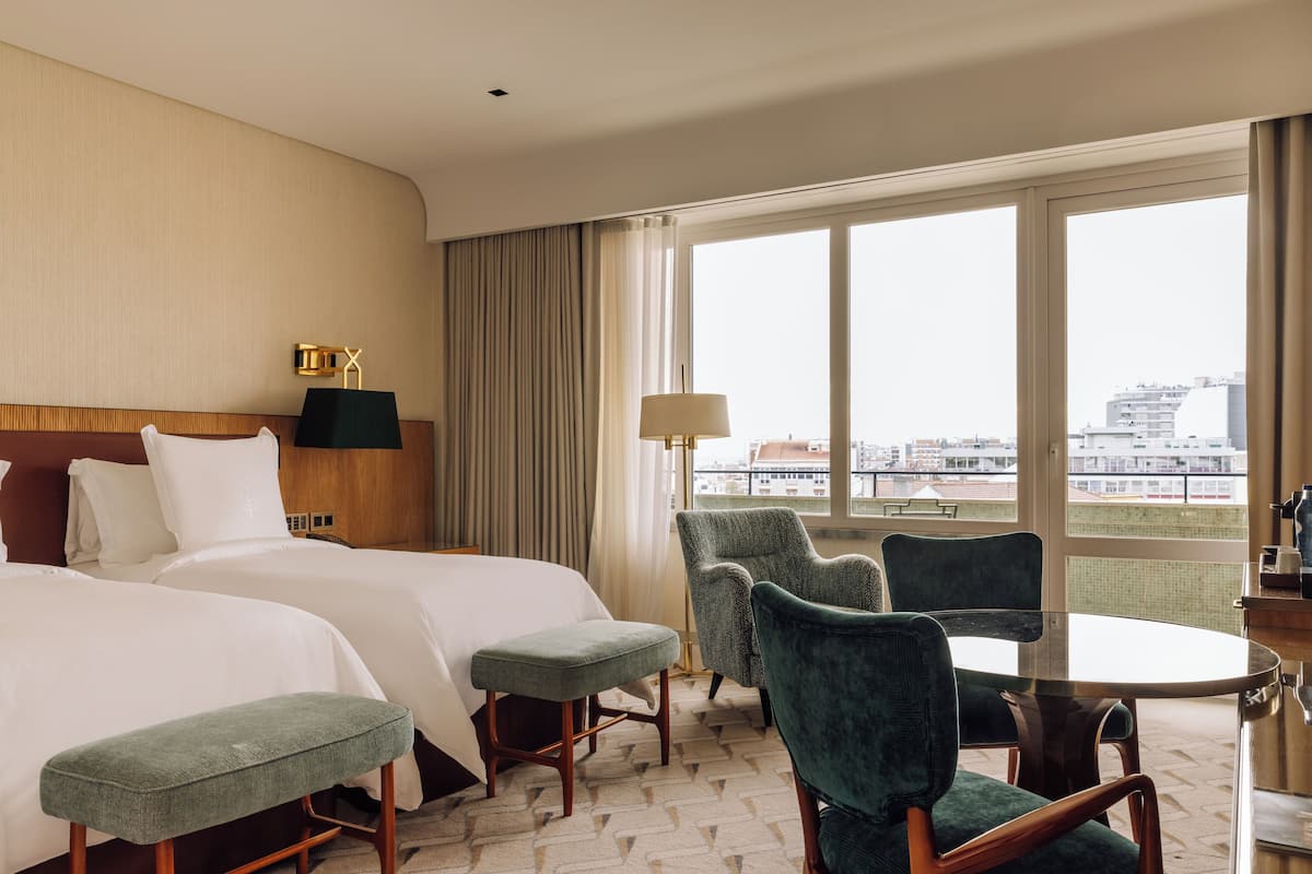A hotel room with two beds, a dining table, green upholstered chairs, and a view of the city through large windows.