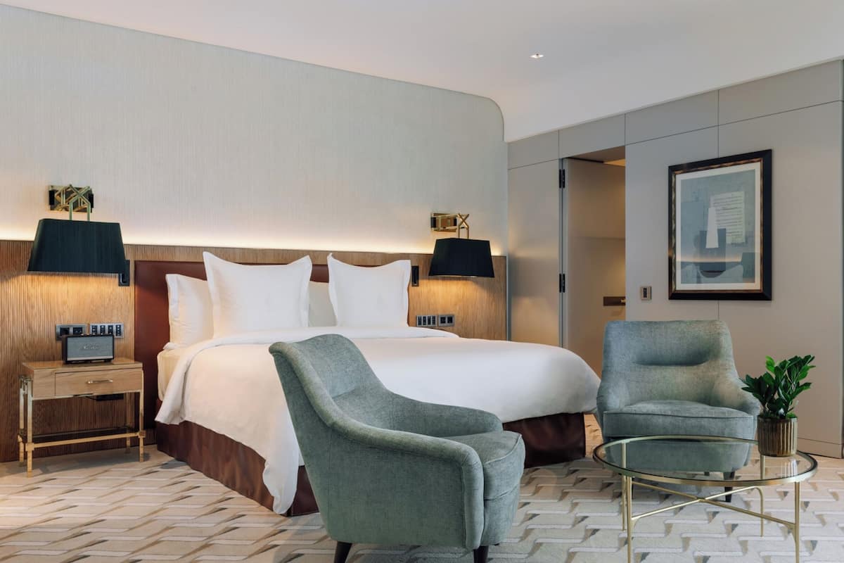 A modern hotel room with a large bed, two bedside tables, a seating area with a chair and a glass coffee table, and a framed picture on the wall.