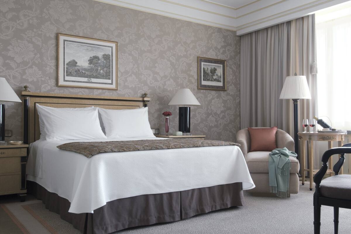 A neatly arranged hotel room with a large bed, a sofa, a chair, and a table with a lamp.