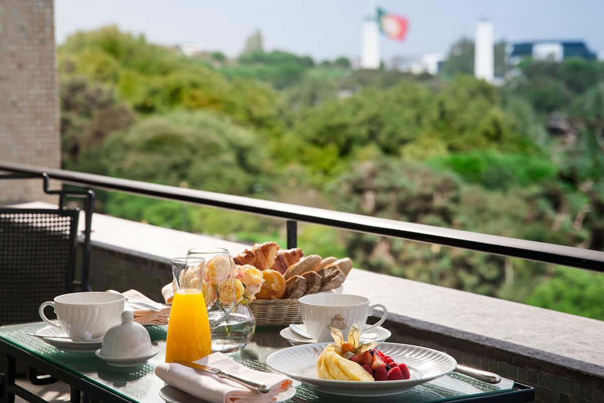 A breakfast table with pastries, fruit, and juice on a balcony overlooking greenery and buildings.