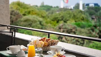 A breakfast table with pastries, fruit, and juice on a balcony overlooking greenery and buildings.