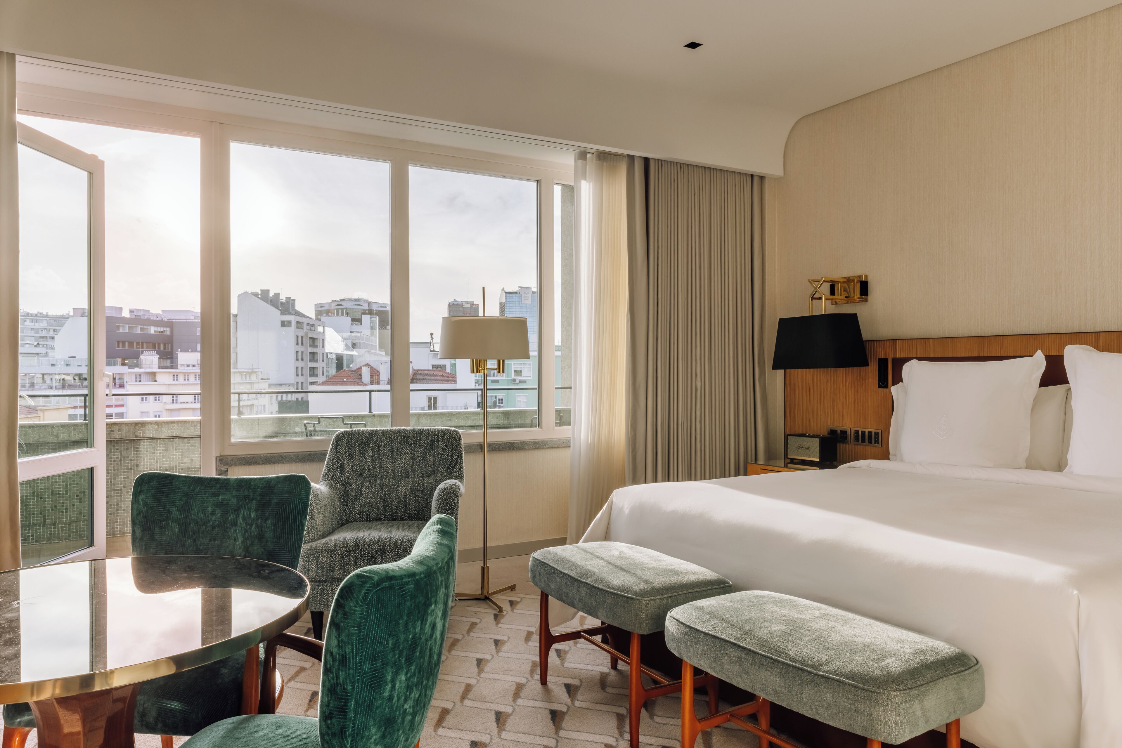 Habitación de hotel con una cama grande, dos sillones verdes, una mesita y vistas a la ciudad a través de amplios ventanales.