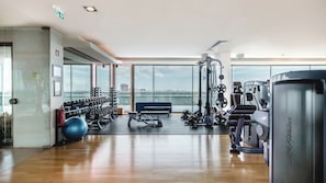 A spacious gym with various exercise equipment, large windows offering a view of a body of water and buildings, and a wooden floor.