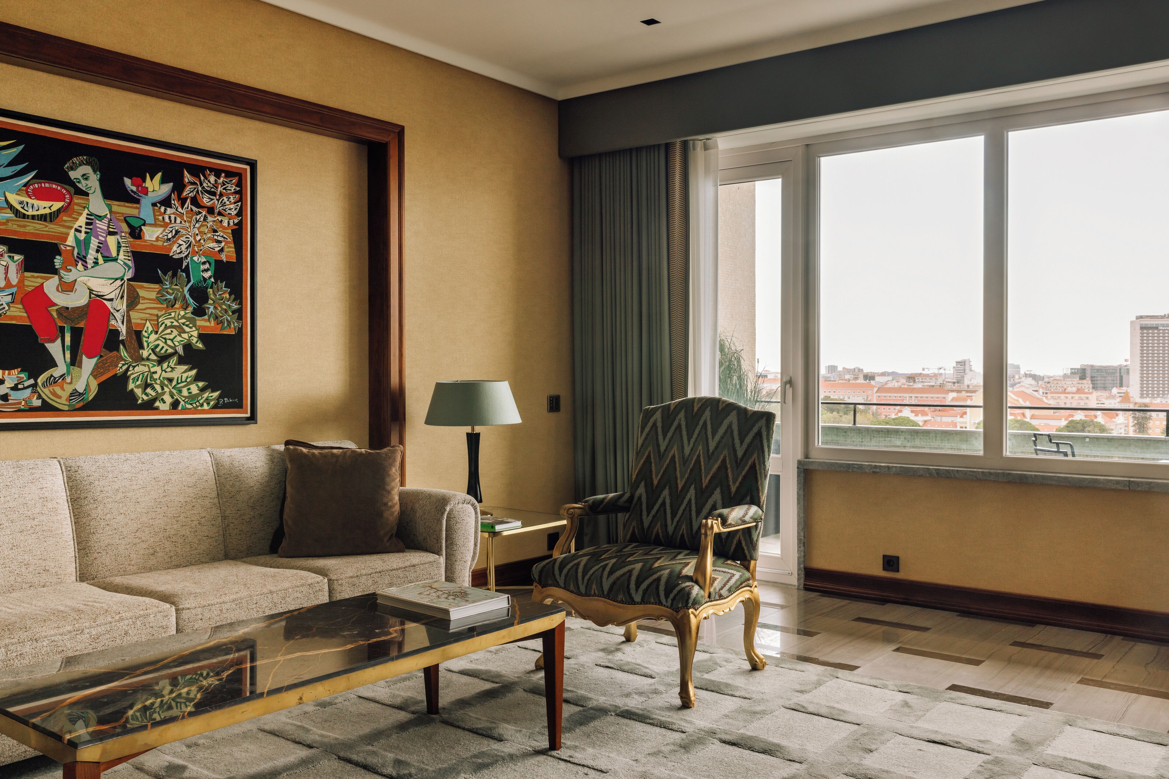 Quarto de hotel com sofá, poltrona e uma grande janela com vista para a cidade.