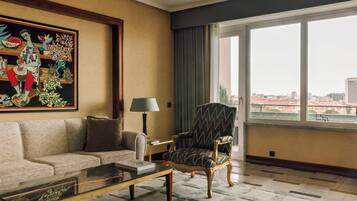 Quarto de hotel com sofá, poltrona e uma grande janela com vista para a cidade.