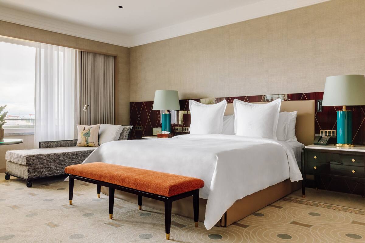 A hotel room with a large bed, two bedside tables, a bench, and a view of the outdoors.
