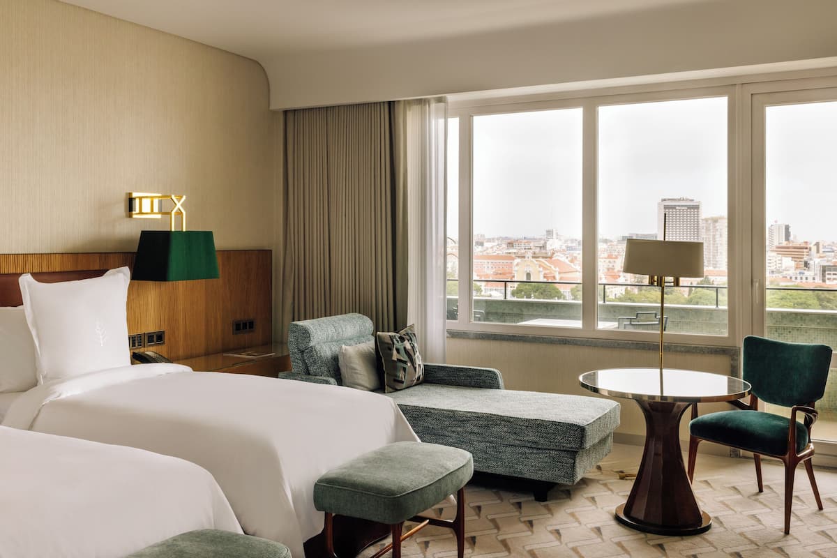 A hotel room with a bed, two armchairs, a small table, and a view of the city through large windows.