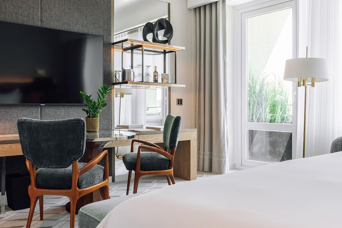 A modern hotel room with a large bed, two armchairs, a desk with a flat-screen TV, a mirror, and a window with curtains.