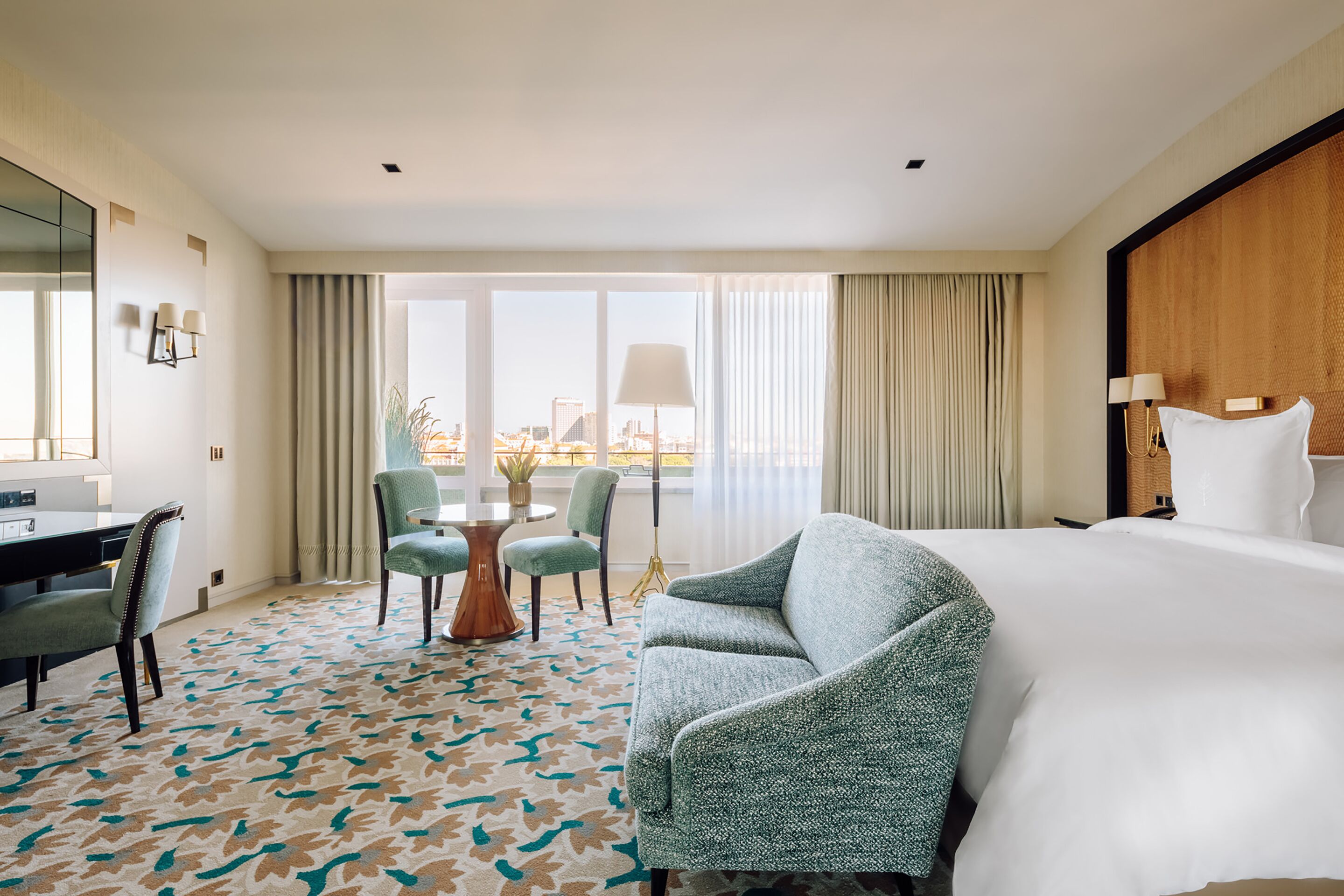 A hotel room with a large bed, a desk with a chair, a small round table with chairs, and a view of the city through large windows.