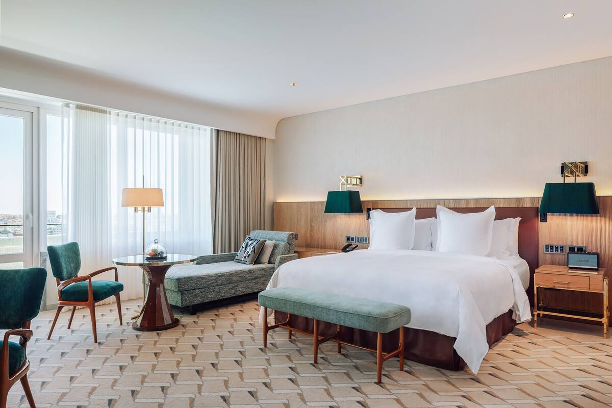 A hotel room with a large bed, a sitting area with a table and chairs, and a view of the city through the window.