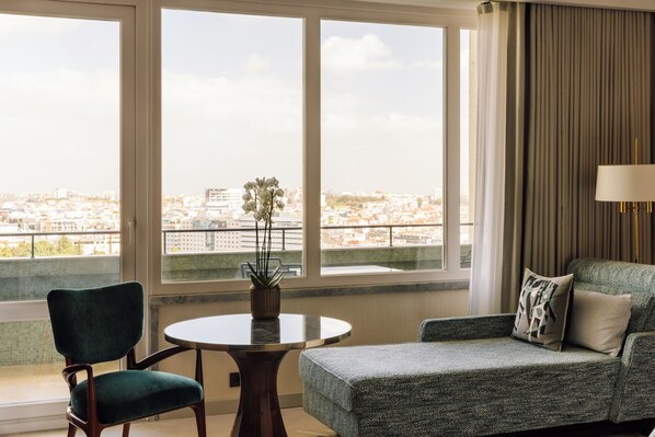 A room with a view of a cityscape, a round table with a potted plant, a green chair, and a grey sofa with a lamp.