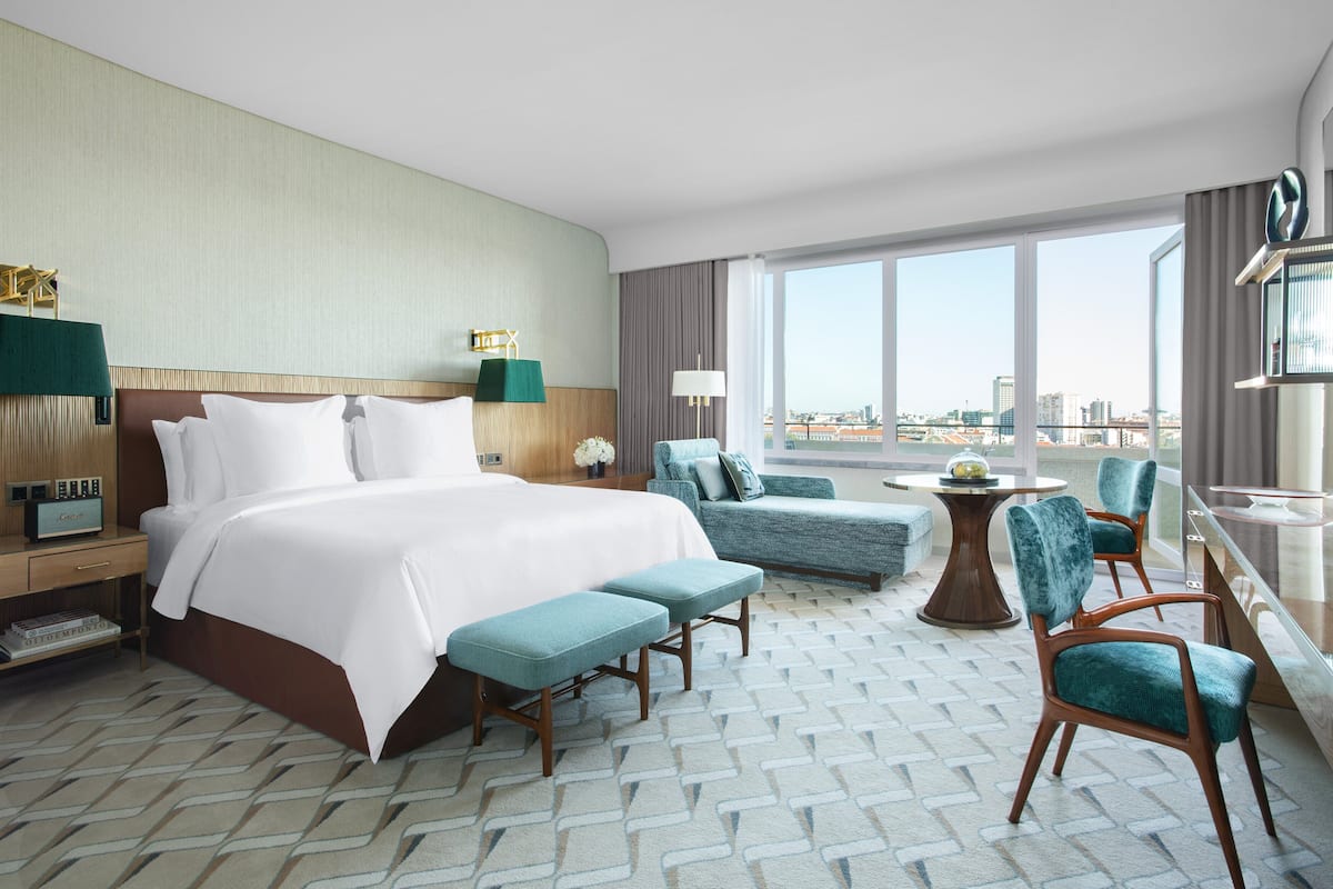 A hotel room with a large bed, two armchairs, a small table, and a view of the city.