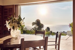 A dining area with a table and chairs, a vase of flowers, and a view of the ocean through a large window.