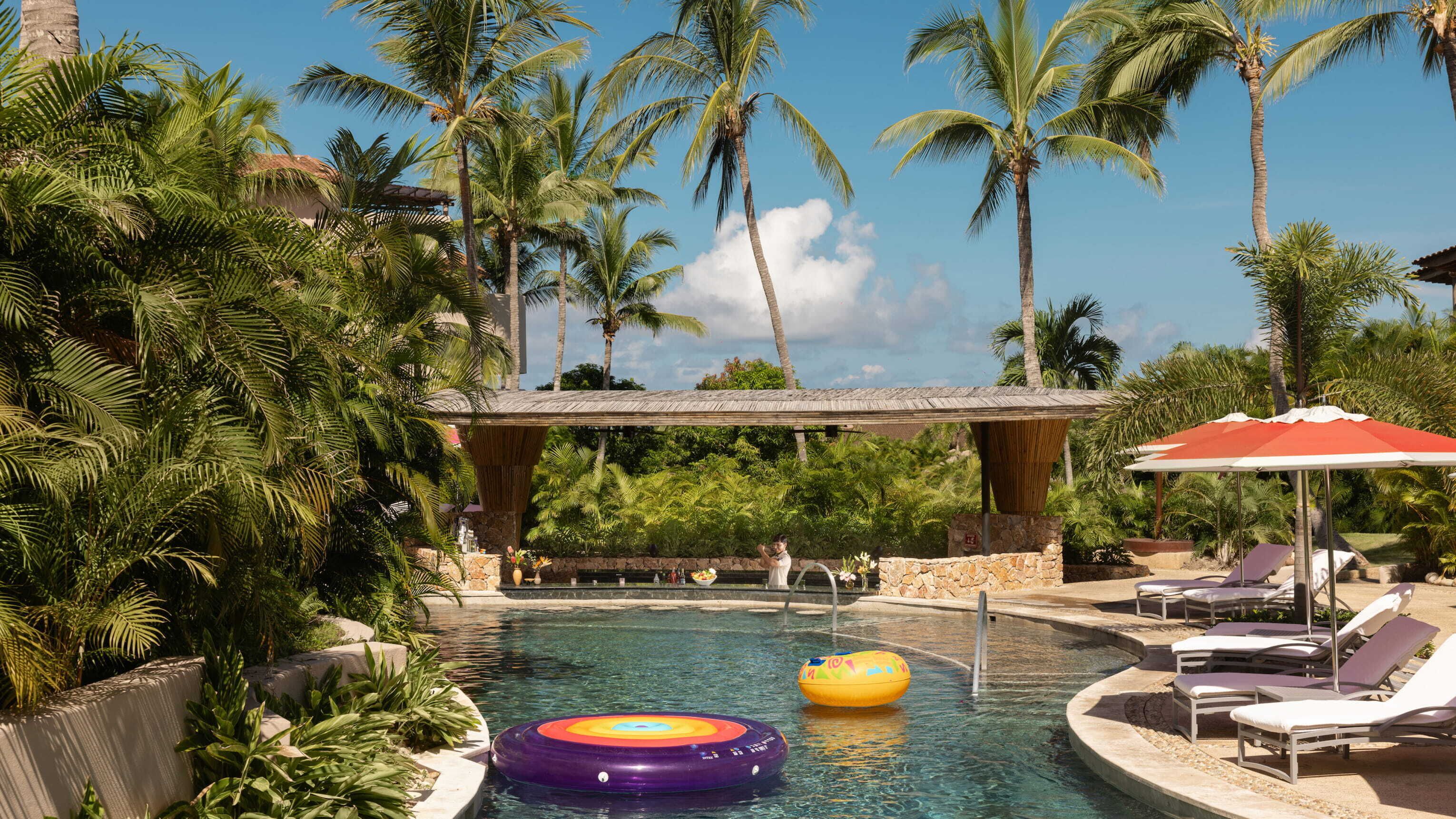 Zona de piscina con sillones de descanso, palmeras y un espacio de estar cubierto.