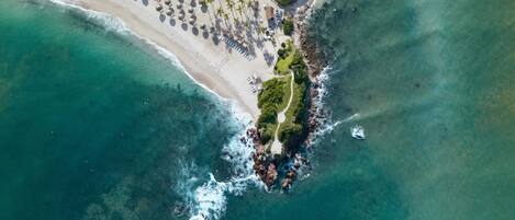 Vista aérea de un complejo turístico en la playa, con arena, sillas de descanso y palmeras.