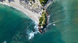 Vista aérea de un complejo turístico en la playa, con arena, sillas de descanso y palmeras.