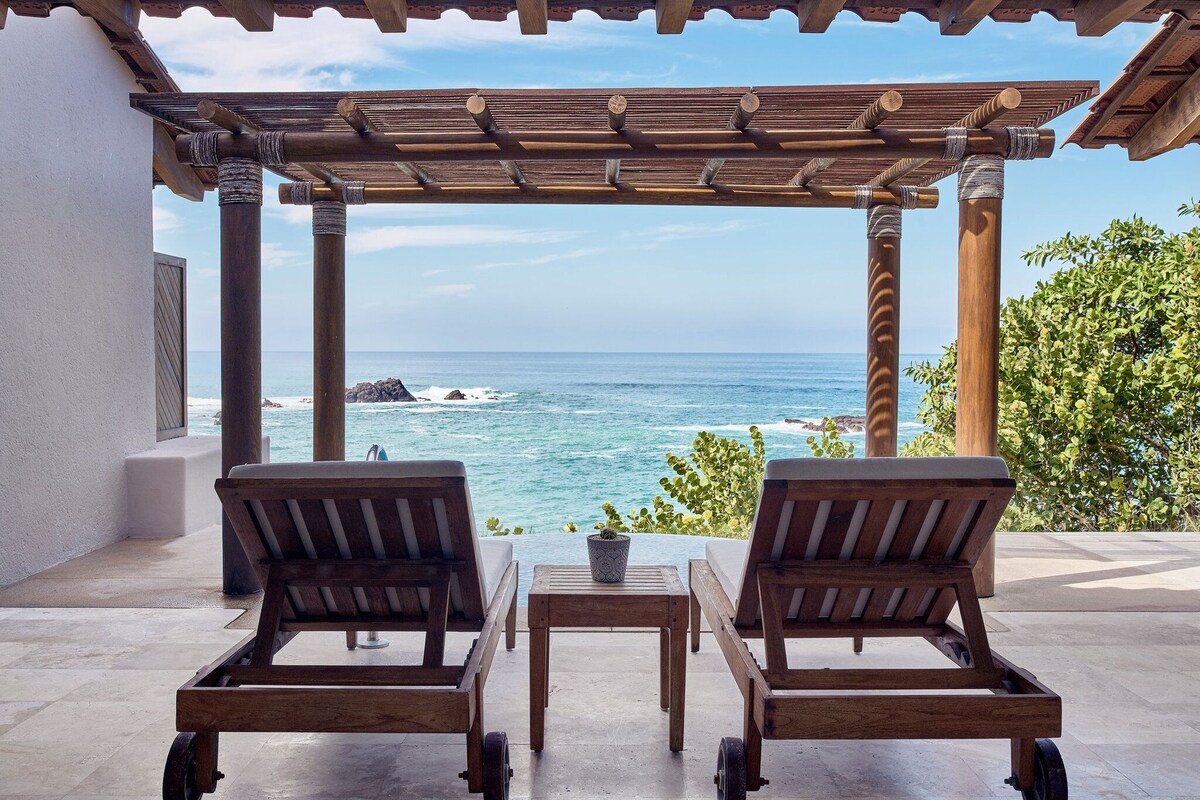 Una pérgola de madera con dos sillones de descanso y una mesita, con vistas a la costa, el océano y el litoral rocoso.