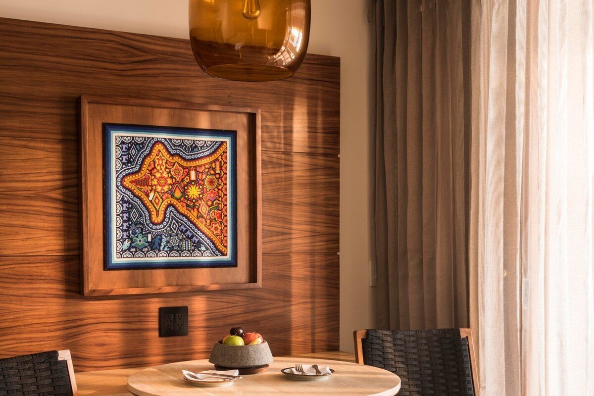 Una habitación moderna con una mesa redonda de madera, dos sillas, una lámpara colgante, una obra de arte enmarcada y una ventana con cortinas.