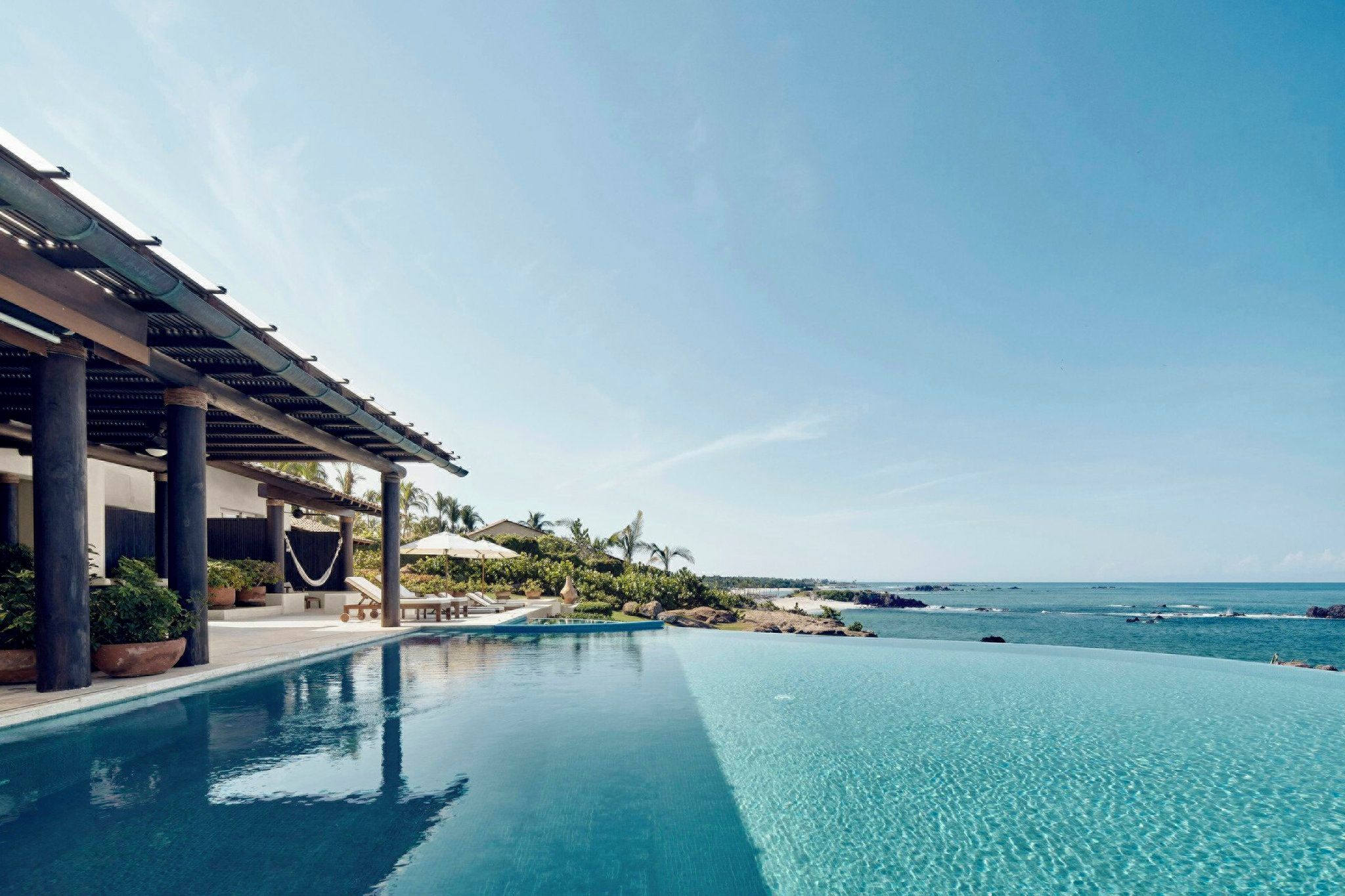 Una piscina infinita azul cristalina con vista al océano y una zona cubierta con sillones de descanso.