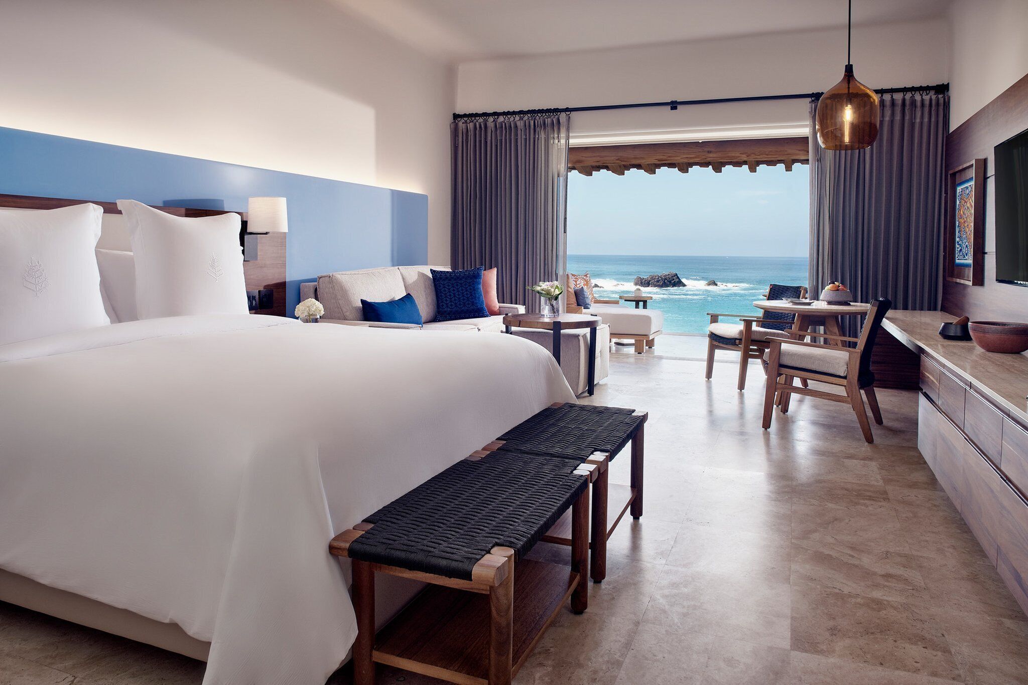 A hotel room with a large bed, a bench, a desk, and a view of the ocean.