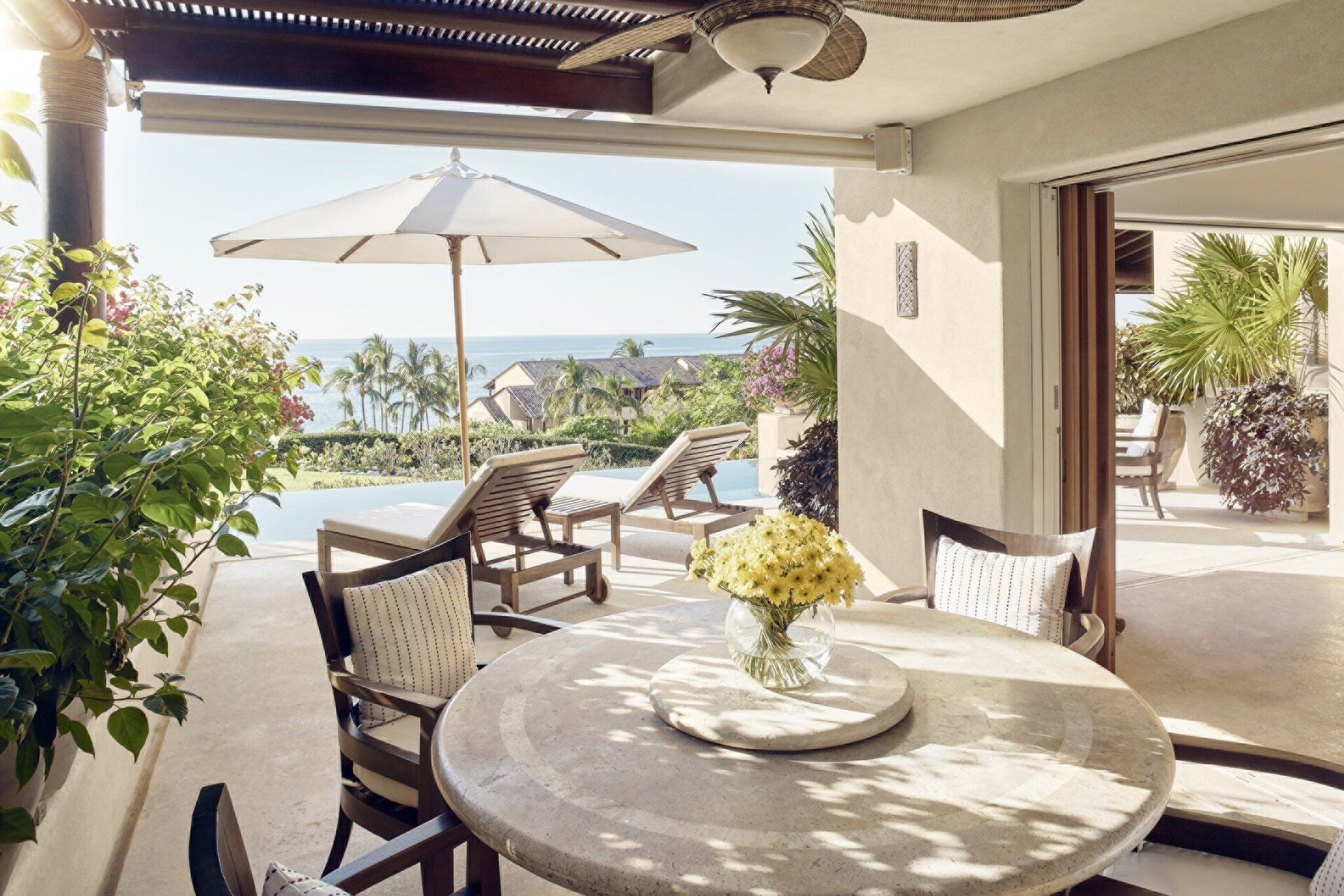 Een terras met een ronde tafel, stoelen en een vaas met gele bloemen. Er is een grote parasol en uitzicht op de oceaan en palmbomen.