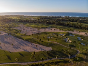 An aerial view of a golf course with sand bunkers, palm trees, and a view of the ocean.