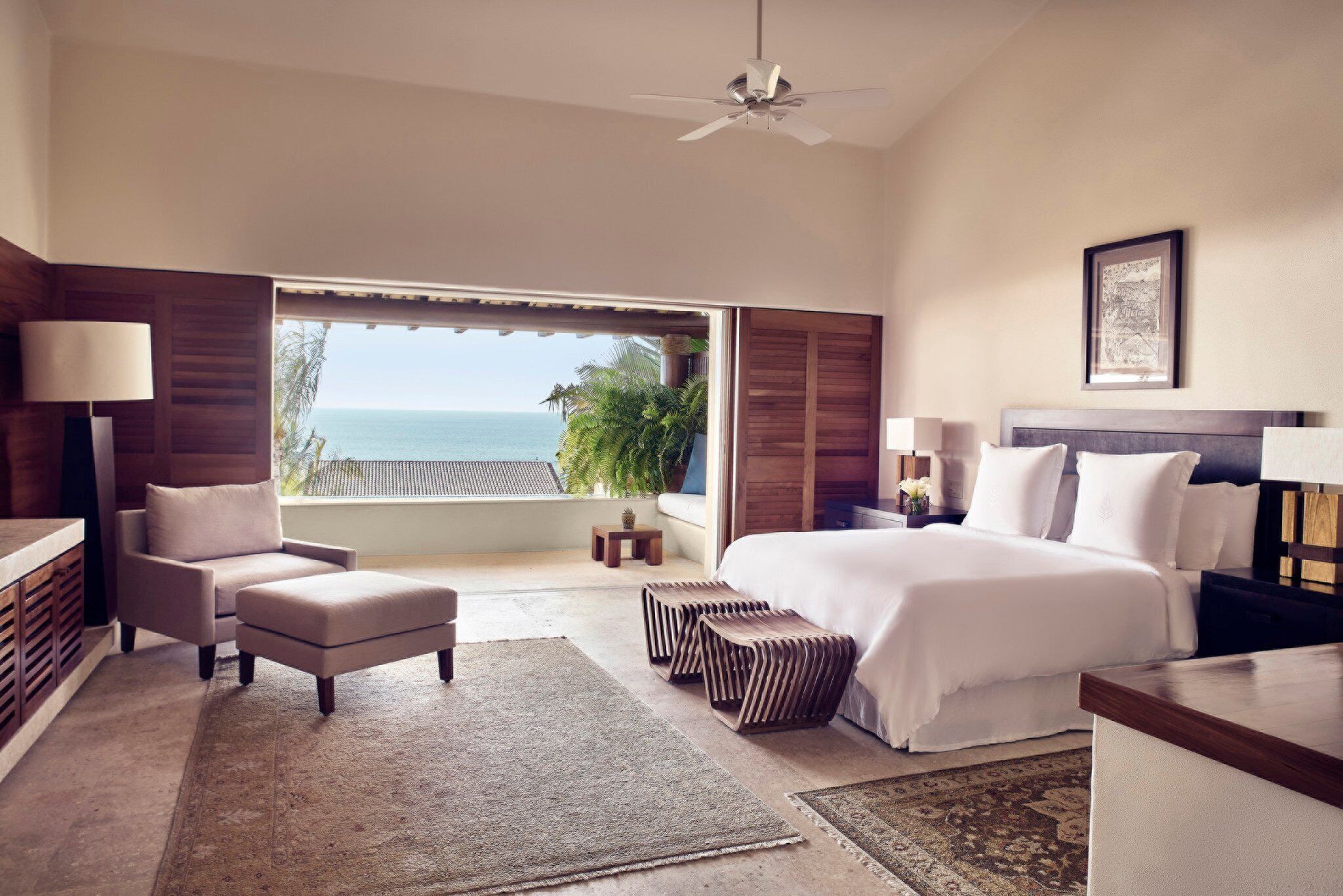 A bedroom with a large bed, a chair, a desk, and a view of the ocean.
