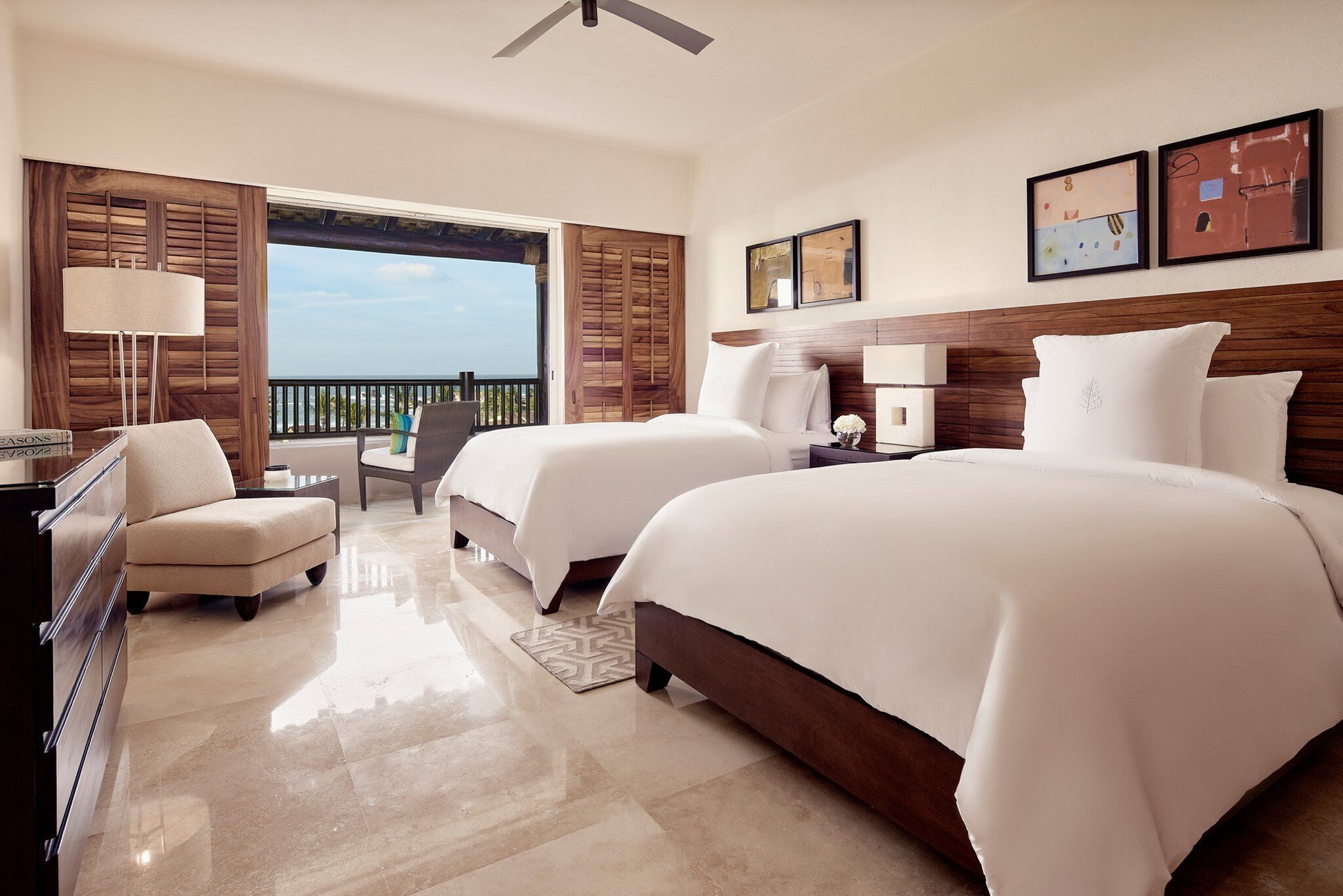 A hotel room with two beds, a desk, a chair, a balcony with a view, and artwork on the wall.